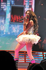 A brunette teenager from a left profile singing into a microphone. She is wearing a short, white tutu dress.