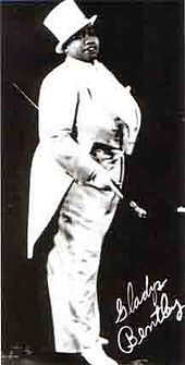 Foto publicitaria de una recia mujer afroamericana vestida con un esmoquin blanco con cola y sombrero de copa, llevando un bastón blanco en la mano y su firma en la esquina inferior derecha