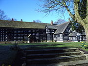 A two-storey black and white timbered building in an L-shape. A wide set of stone steps in the foreground leads onto a grassed area in front of the house.