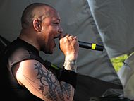 Un hombre, con un gran tatuaje en su brazo derecho, canta en un micrófono mientras mira a la audiencia.