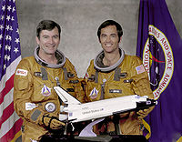 Los miembros de la tripulación John W. Young y Robert L. Crippen posan con el traje de eyección (EES) con un pequeño modelo del transbordador Espacial.