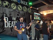 A color photograph of three young men playing on the The Bealtes: Rock Band instruments in front of a large display for the game.