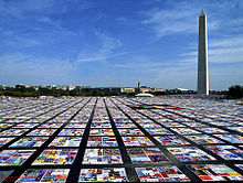 Squares of a quilt laid out in sections in a grid-like pattern, on a large, flat paved surface. The Washington Monument, a tall obelisk, can be seen in the background.