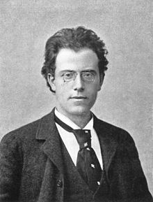  A young man wearing rimless spectacles looks straight out of the picture. He is formally dressed in dark jacket, white shirt and loose necktie. His longish hair is brushed back to reveal a wide forehead.