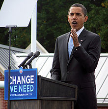 Obama gestures from the podium while campaigning. The front of the podium has a sign that reads "Change We Need" with WWW. BARACKOBAMA. COM below and his campaign logo above.