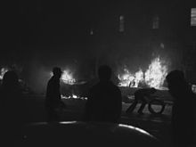People silhouetted against cars in the background, which are engulfed in flames. One person is bent over and holding a doughnut-shaped object.