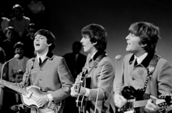 Una imagen de negro y blanco de tres hombres a tocar la guitarra. Llevan gris abotonado chaquetas con lazos por debajo. Una audiencia es visible detrás de ellos a la izquierda.